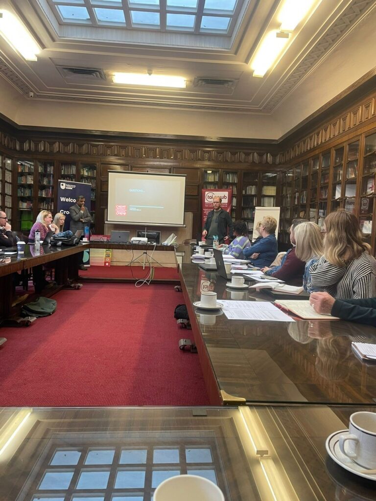 Rob Milligan stood at the front of a red carpeted room with wooden bookshelves surrounding, presenting to people sat at glass tables listening