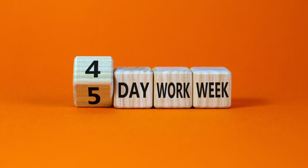 Four wooden blocks that say '4 day work week.' The block with 4 on it is in motion and the 5 can be seen written on the underside of the block